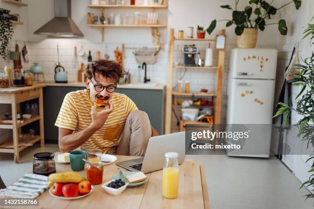 jeune homme habillé de façon décontractée utilisant un ordinateur portable tout en prenant un petit-déjeuner - tartine photos et images de collection