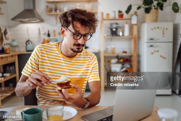 jeune homme vêtu avec désinvolture utilisant un ordinateur portable tout en prenant un petit-déjeuner - tartine photos et images de collection