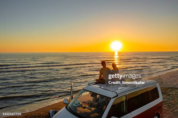 pareja viendo la puesta de sol mientras está sentada en el techo de la autocaravana en la playa - viaje por carretera fotografías e imágenes de stock