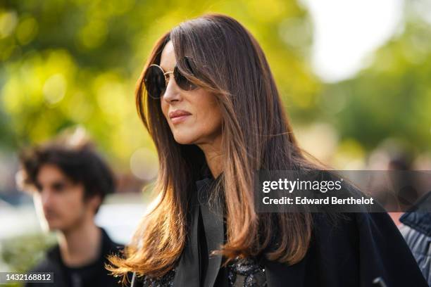 Monica Bellucci wears black circle sunglasses, a black tulle with embroidered sequined flower pattern shirt, a black silk knot tie, a black long...