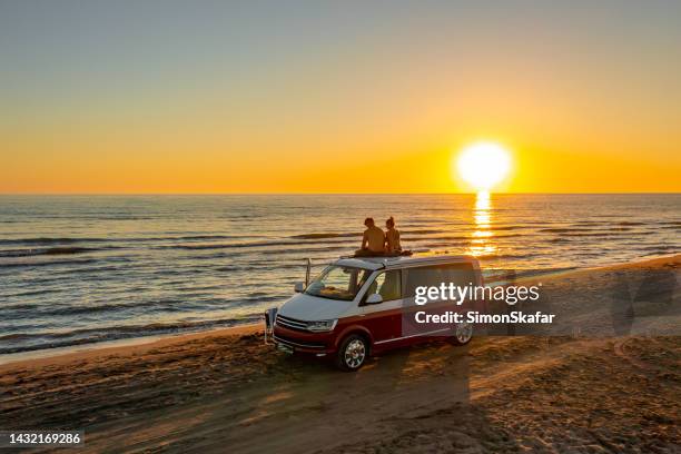pareja sentada en el techo de la autocaravana en la playa mientras disfruta de la puesta de sol - trailer beach fotografías e imágenes de stock