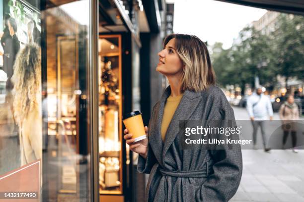 beautiful woman with a cup of coffee stands in front of a store window in the city. - window shopping stock pictures, royalty-free photos & images