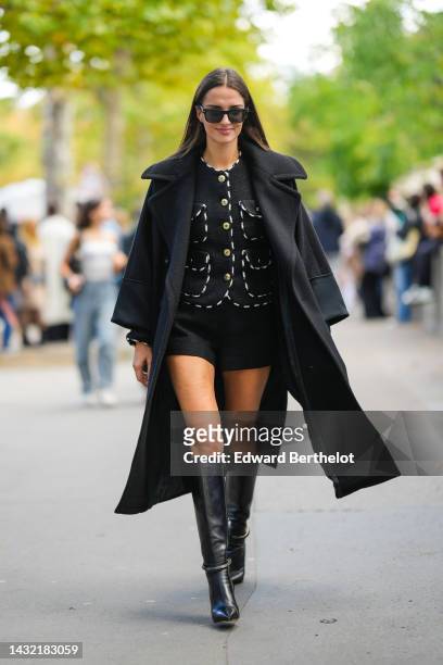 Gabrielle Caunesil wears black sunglasses, a black tweed / white borders / gold buttons jacket, a black oversized long coat, black shorts, black...