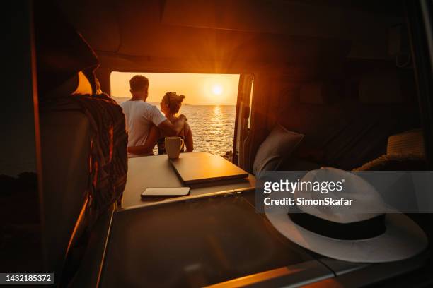 paar genießt den sonnenuntergang über dem meer, während es zusammen vor dem wohnmobilanhänger steht - caravan stock-fotos und bilder