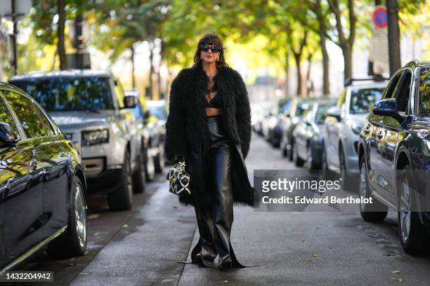 Gili Biegun wears black sunglasses, gold earrings, a black V-neck / cropped bra underwear, a black curly fur oversized long coat, a black shiny...