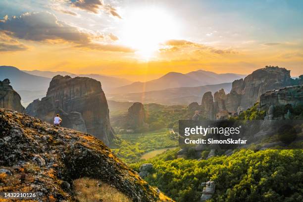 tourist exploring meteora monastery while sitting on rock against orange sky - meteora greece stock pictures, royalty-free photos & images