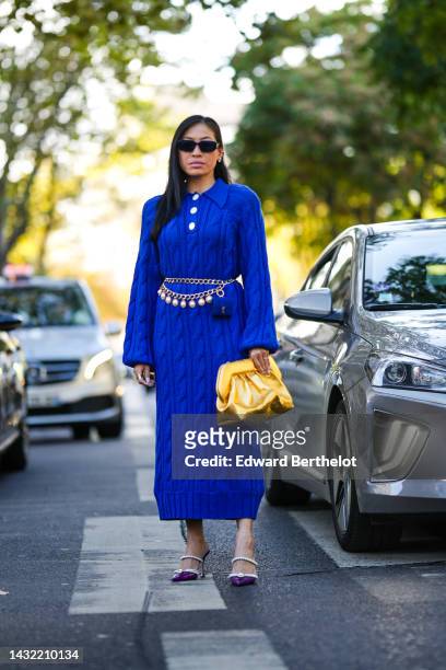 Miki Cheung wears black sunglasses, earrings, a royal blue embroidered wool / white buttoned long pullover dress, a gold pendants belt, a gold shiny...