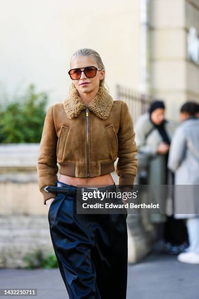 Caroline Daur wears black and brown sunglasses, a brown suede with sheep collar cropped jacket, a black shiny leather belt, black large wide legs...