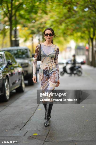 Mary Leest wears black square sunglasses, silver earrings, a pale blue / yellow / brown print pattern transparent long sleeves / long tube dress, a...