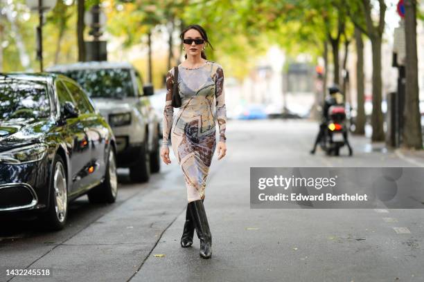 Mary Leest wears black square sunglasses, silver earrings, a pale blue / yellow / brown print pattern transparent long sleeves / long tube dress, a...