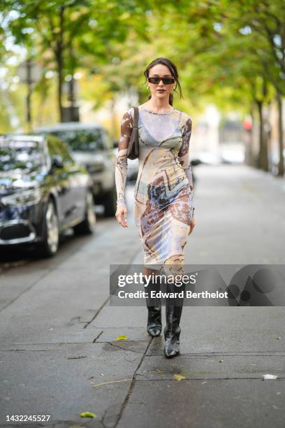 Mary Leest wears black square sunglasses, silver earrings, a pale blue / yellow / brown print pattern transparent long sleeves / long tube dress, a...