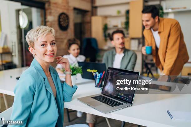 smiling woman with blond short hair sitting in the office and looking directly at the camera, while her colleagues are seen in the background - java stock pictures, royalty-free photos & images