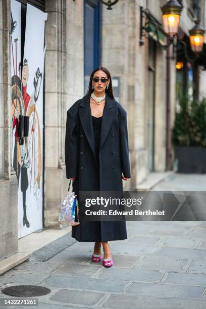 Elisa Taviti wears black cat eyes sunglasses, silver large earrings, a white pearls large necklace, a black V-neck long dress, a black buttoned long...