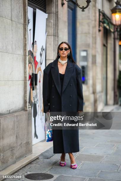 Elisa Taviti wears black cat eyes sunglasses, silver large earrings, a white pearls large necklace, a black V-neck long dress, a black buttoned long...