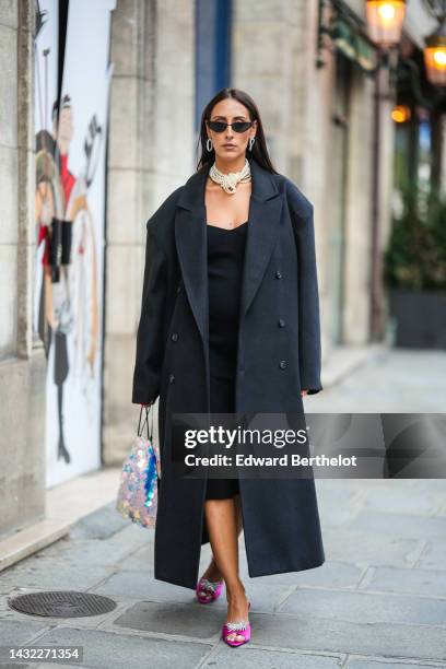Elisa Taviti wears black cat eyes sunglasses, silver large earrings, a white pearls large necklace, a black V-neck long dress, a black buttoned long...