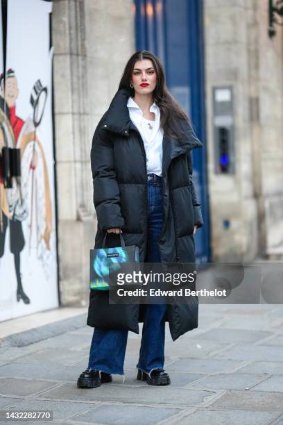 Zoia Mossour wears silver earrings, a white tank-top, a silver long chain necklace, a white shirt, a black oversized long puffy coat, a black with...