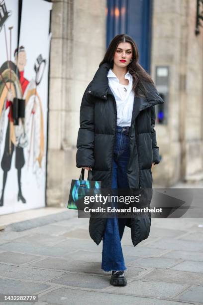 Zoia Mossour wears silver earrings, a white tank-top, a silver long chain necklace, a white shirt, a black oversized long puffy coat, a black with...