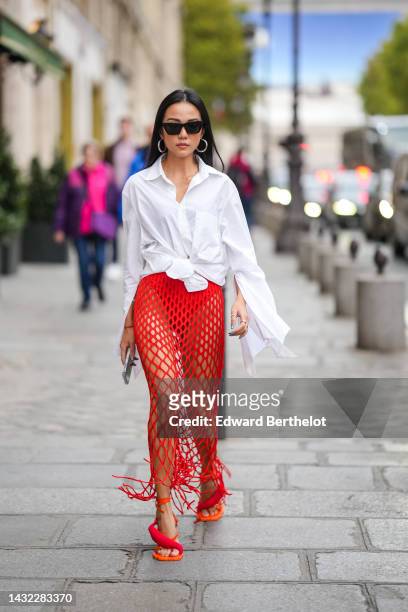 Yoyo Cao wears black sunglasses, silver earrings, a white oversized shirt, a red mesh / fishnet long fringed skirt, silver rings, orange shiny...