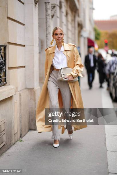 Leonie Hanne wears gold hearts pendants earrings, a white cropped shirt, a gold shiny silk long coat, a beige matte leather handbag, gold rings, a...