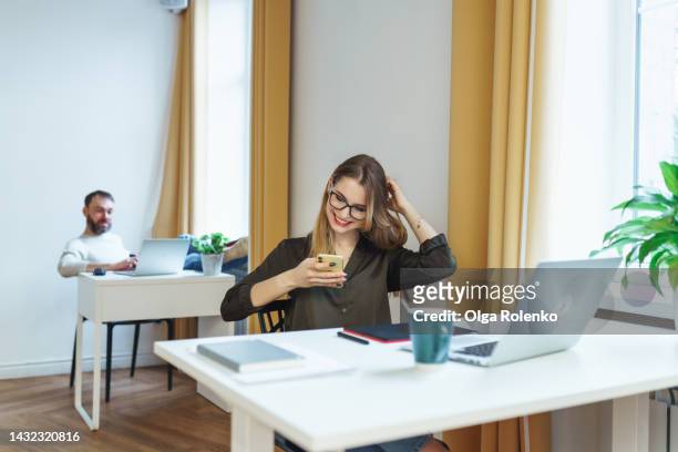 flirt chatting at work. smiling woman in eyeglasses using smartphone at work against male colleague at desk - romance de oficina fotografías e imágenes de stock