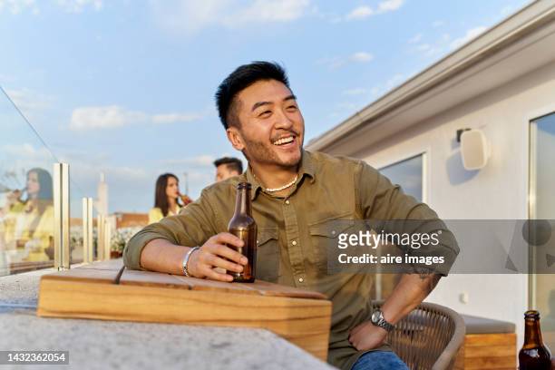 a smiling asian man with a beard and a shirt holding a beer and behind him other people - hand holding a bottle stock pictures, royalty-free photos & images