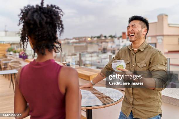 a bearded asian man smiling and holding a gin and tonic in his hand and in front of him a colored woman with curly hair - gin stockfoto's en -beelden