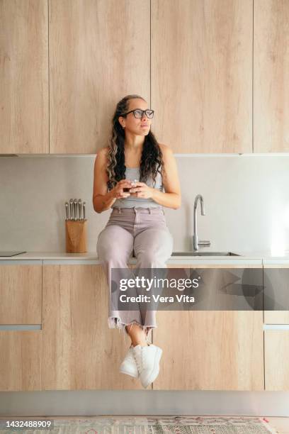 vertical front view of hispanic woman with coffee at home sit on countertop kitchen. - vita domestica fotografías e imágenes de stock