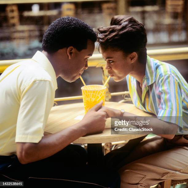 1980s Romantic Couple Boy And Girl Face To Face Holding Hands Sharing A Soft Drink With Two Straws.