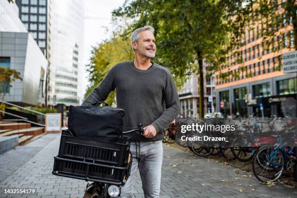 handsome mature man riding cargo bicycle and contributes to eco-friendly environment. businessman near green buildings - dutch culture stock pictures, royalty-free photos & images