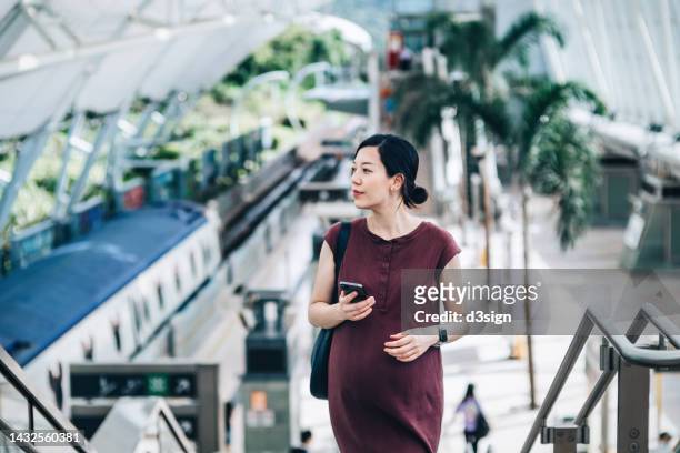 young asian pregnant businesswoman using smartphone while commuting with subway in the city, looking sideways while walking upstairs in train platform. pregnant woman commuting to work - metro platform stockfoto's en -beelden