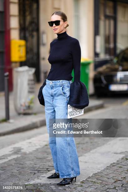 Guest wears black futurist sunglasses, gold earrings, a black turtleneck pullover with large puffy flared sleeves, blue faded denim wide legs pants,...