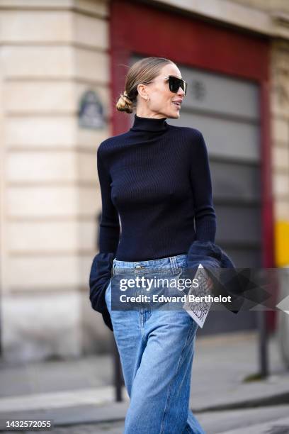 Guest wears black futurist sunglasses, gold earrings, a black turtleneck pullover with large puffy flared sleeves, blue faded denim wide legs pants,...