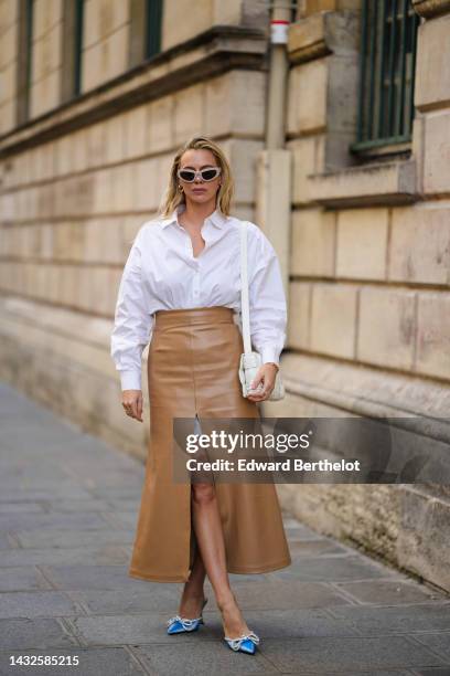 Guest wears beige cat eyes sunglasses, silver earrings, a white shirt, a brown shiny leather slit / split long skirt, a white shiny leather shoulder...