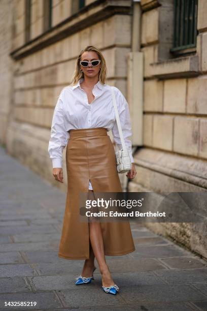 Guest wears beige cat eyes sunglasses, silver earrings, a white shirt, a brown shiny leather slit / split long skirt, a white shiny leather shoulder...