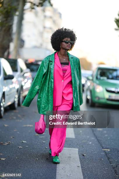 Guest wears black sunglasses, gold earrings, a gold chain necklace, a neon pink with gold and silver print pattern shoulder-off top, a neon pink...