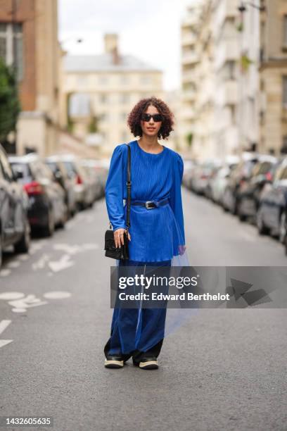 Guest wears black sunglasses, a royal blue pleated / accordion silk blouse, a black shiny leather with silver buckle belt, a black shiny leather...