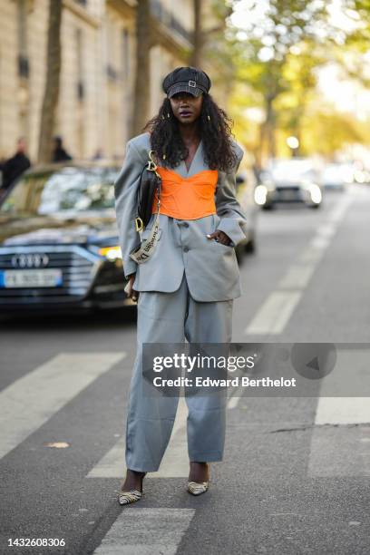 Guest wears a black with white seams cap, gold hair clips, a gray oversized blazer jacket, a neon orange corset, pale gray suit pants, a black shiny...