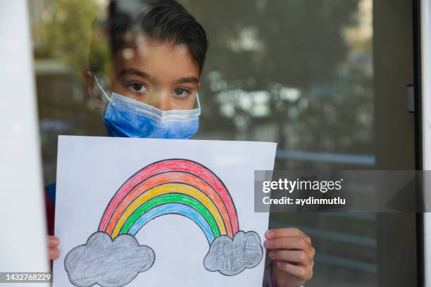 kleiner junge mit gesichtsmaske schaut durch das fenster und zeigt ihre zeichnung zu hause während der pandemie - rainbow kids stock-fotos und bilder