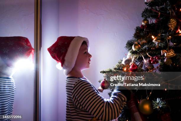 little boy in christmas hat near christmas tree at home. neon light, evening. - christmas tree presents stock pictures, royalty-free photos & images