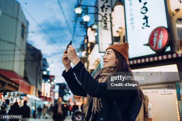 smiling young asian female traveller photographing the vitality and colourful neon signboards of busy downtown city street scene with smartphone while travelling and exploring in osaka, japan. travel, vacation and holiday concept - stadt osaka stock-fotos und bilder