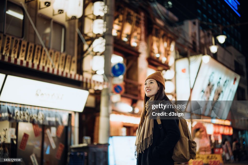 Young Asian female traveller with backpack visiting Japan, exploring and strolling in downtown city street at night in Osaka. A popular nightlife and entertainment area in the city. Travel, vacation and holiday concept