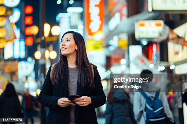 young asian female traveller with backpack searching for directions with smartphone gps navigation app while exploring and strolling in busy downtown city street at night in osaka, japan. lifestyle and technology. travel, vacation and holiday concept - japanese woman looking up stockfoto's en -beelden