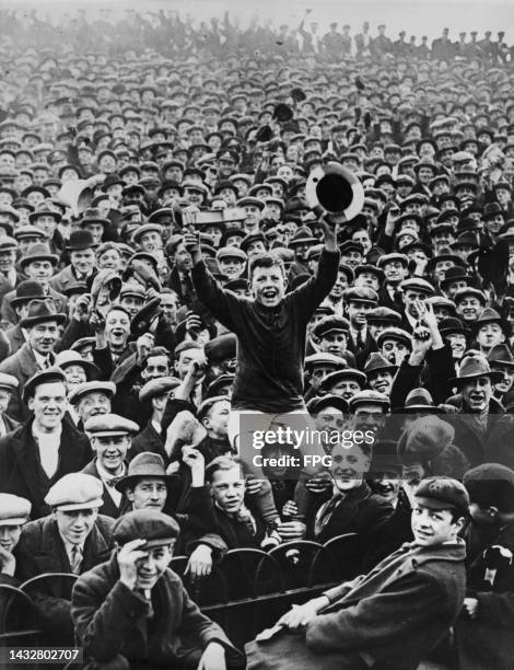 Young boy, waving a football rattle and a hat as he sits on the shoulders of a man in a crowd of, possibly, football supporters, attending a match,...