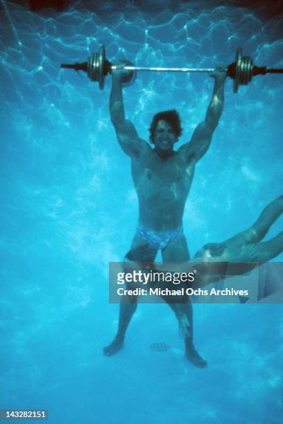 Austrian Bodybuilder Arnold Schwarzenegger lifts a barbell at the bottom of a pool as a topless girl swims by circa 1977 in Los Angeles, California.