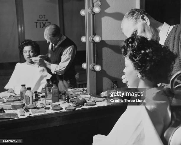 Make-up artist applying lipstick to a woman, draped in a protective sheet, sitting looking at her reflection in her dressing room mirror, United...