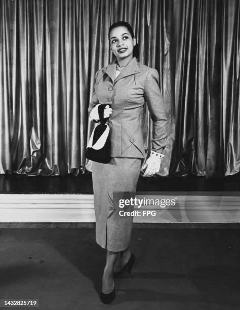 Woman poses in a grey suit with a mid-length skirt, with white gloves and holding a black-and-white purse, with a curtains in the background beyond...