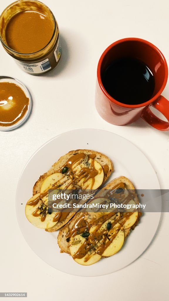 Sweet breakfast, white bread toast with vegan almond cream and apple slices. Raw seed topping. American coffee in red cup. Aerial view on white table.