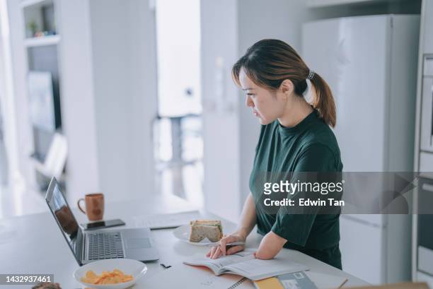 asian chinese woman with disability deformed arm e learning at kitchen counter using laptop while eating sandwiches - amputee woman imagens e fotografias de stock