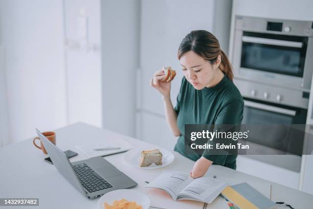 asian chinese woman with disability deformed arm studying at kitchen counter using laptop while eating sandwiches - autonomous stock pictures, royalty-free photos & images