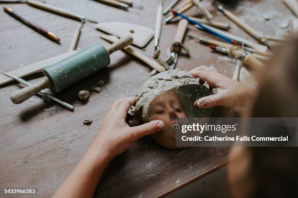 a curious child notices her reflection in a scraper carving tool as she plays with clay - schnitzen stock-fotos und bilder